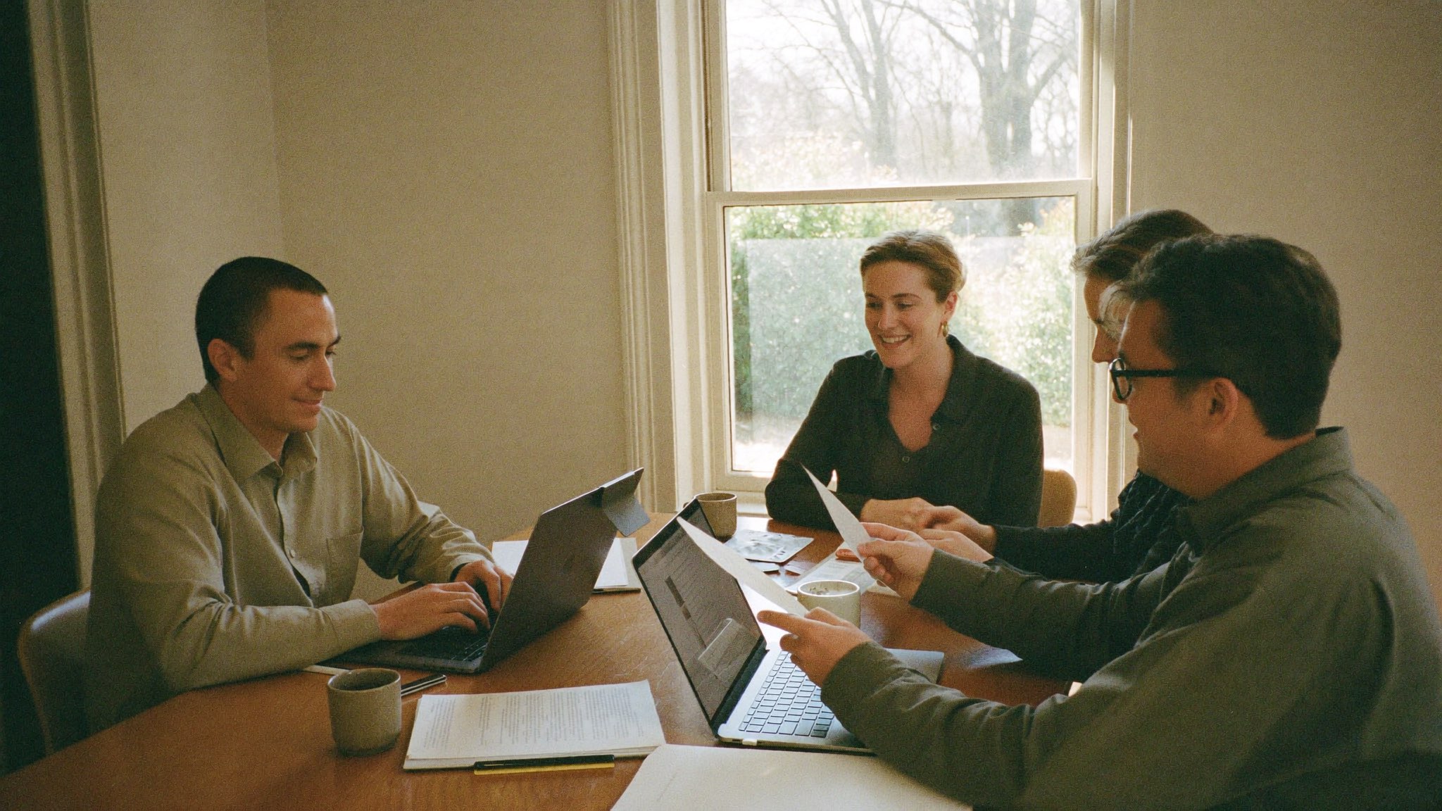 Four people working together around a table in daylight, laptops and printed pages between them